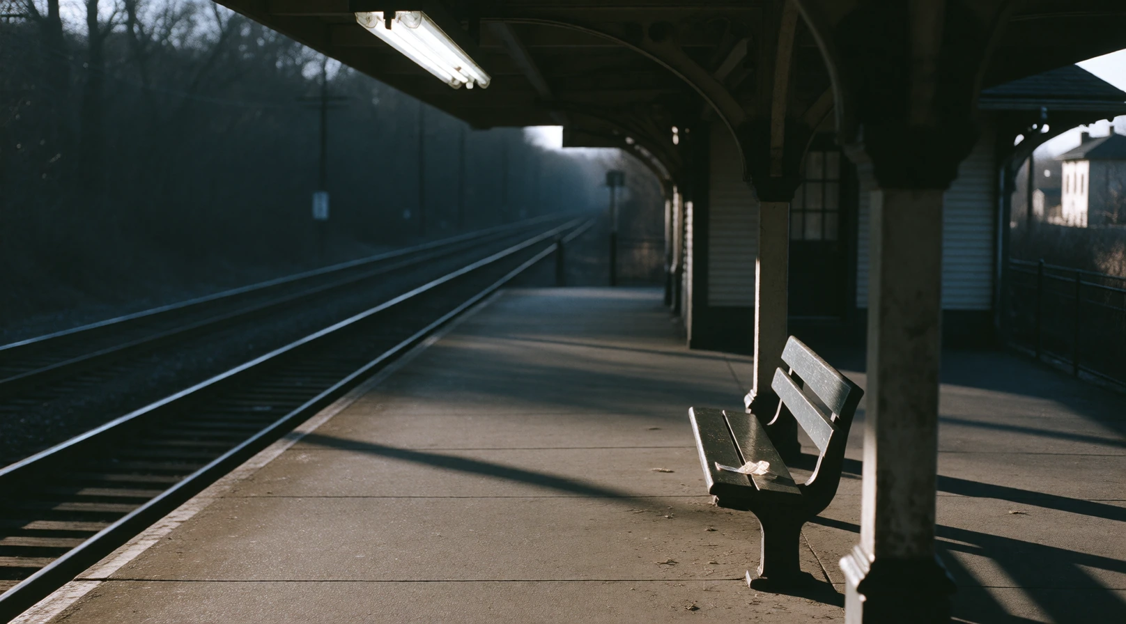 Empty railway platform at dusk, single fluorescent light humming overhead, casting hard shadows across the concrete. A lone bench, a discarded ticket 