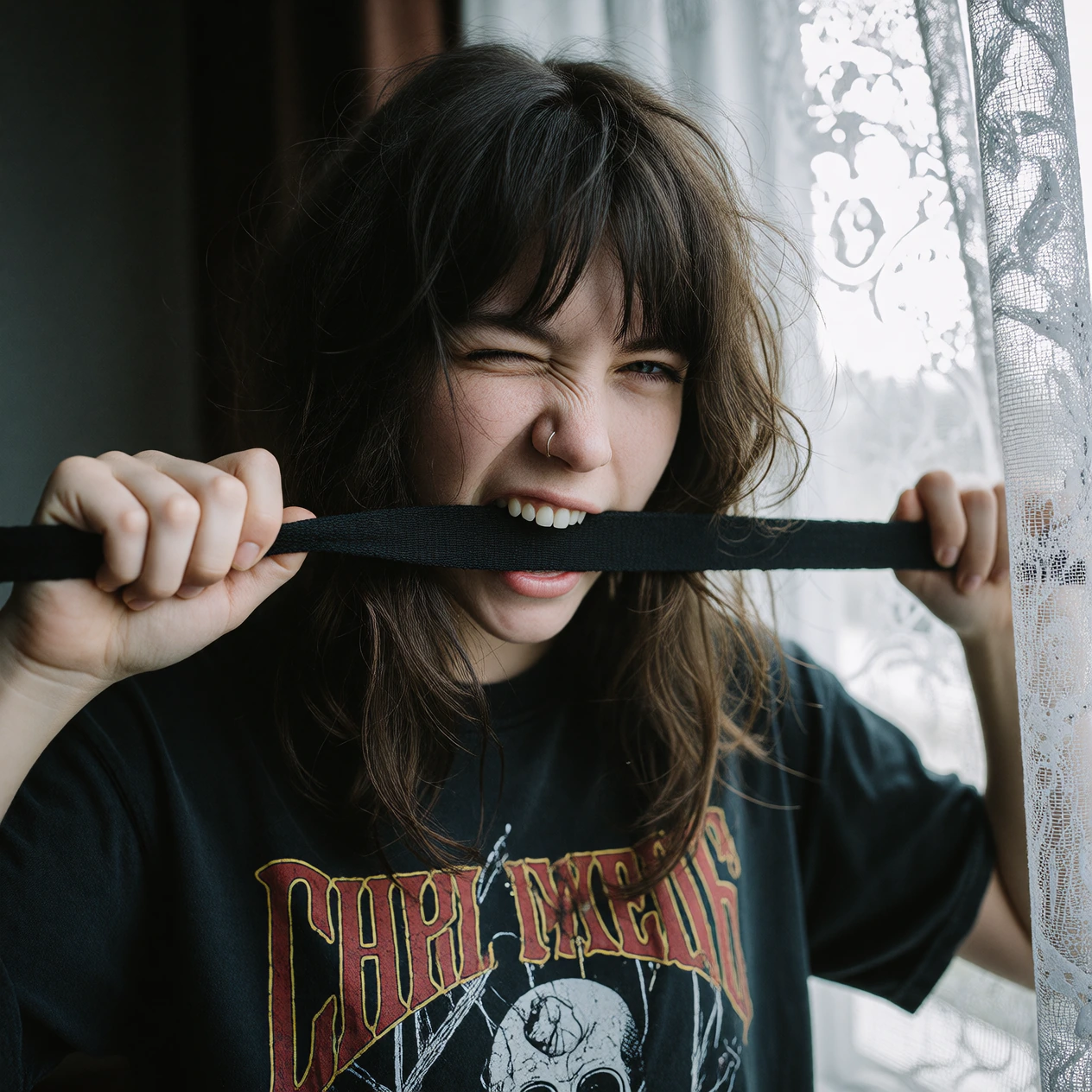 A professional midshot analog photograph of a moody 20yearold woman with a messy brunette fringe haircut, winking one eye while playfully biting down on a long black fabric strap held tightly in both hands. She is wearing a dark vintage graphic band