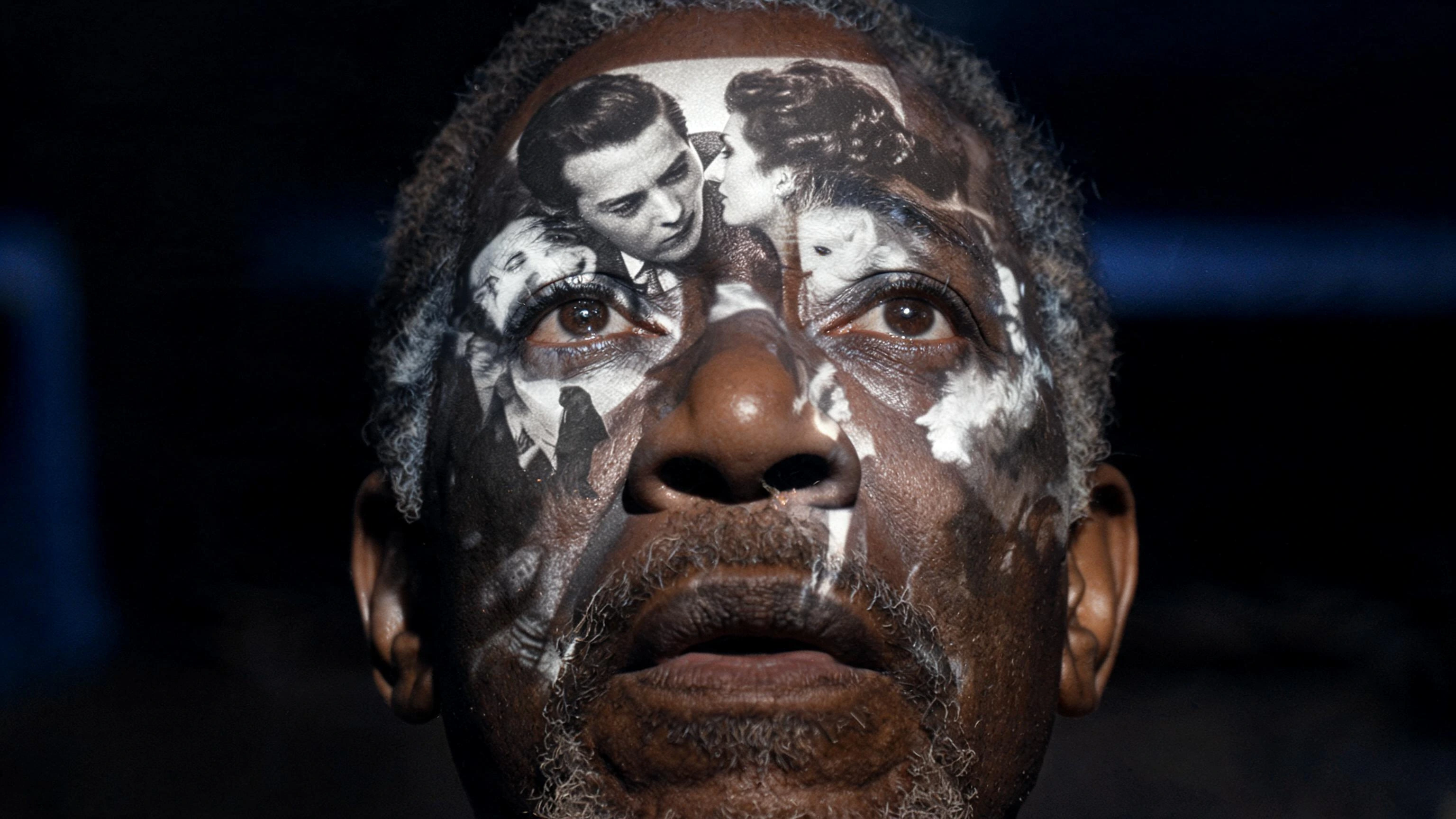 Cinematic keyframe of an older Black man watching an old film. Shot from a low symmetrical angle looking up at his face. He has a deeply emotional, awestruck expression as he watches the projection in front of him. A black and white 1940s Hollywood f