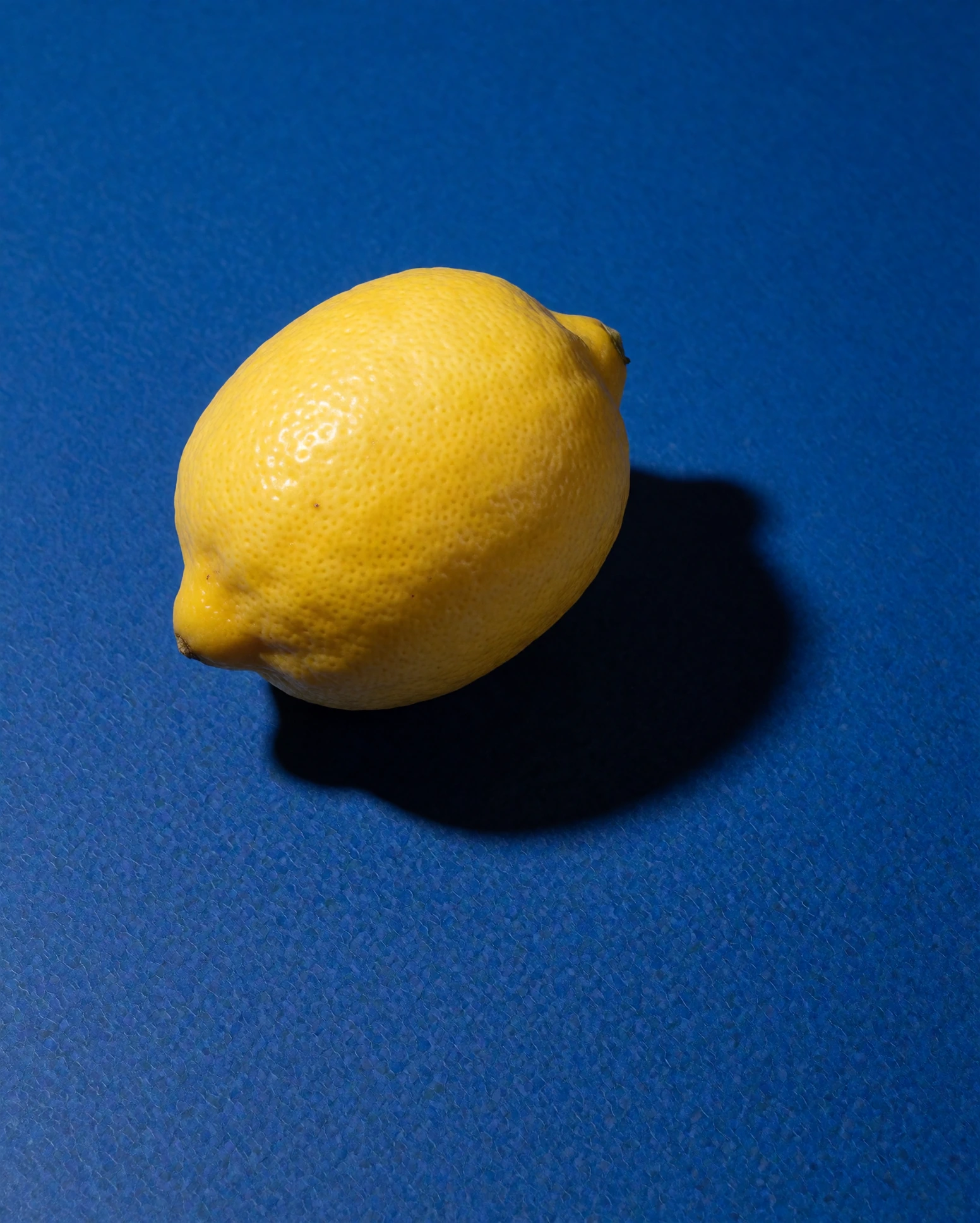 One lemon on a flat cobaltblue surface, hard directional light, crisp shadow. Topdown. That's the whole image. Colorblocked still life, matte finish.