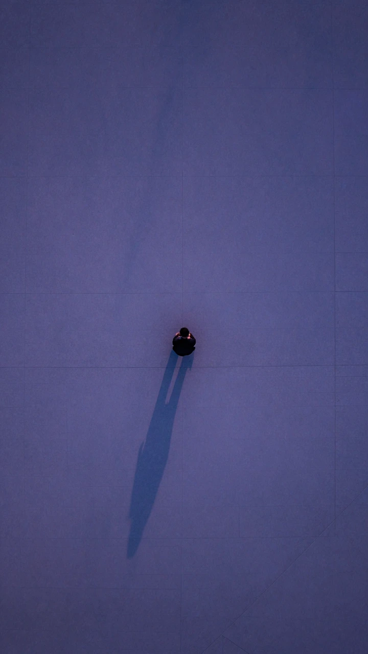Aerial view of a solitary figure sitting on an empty concrete plaza at dusk, shot from directly overhead, their small shadow stretching impossibly long across bare gray stone, surrounded by vast negative space and geometric emptiness, cool blue-purpl