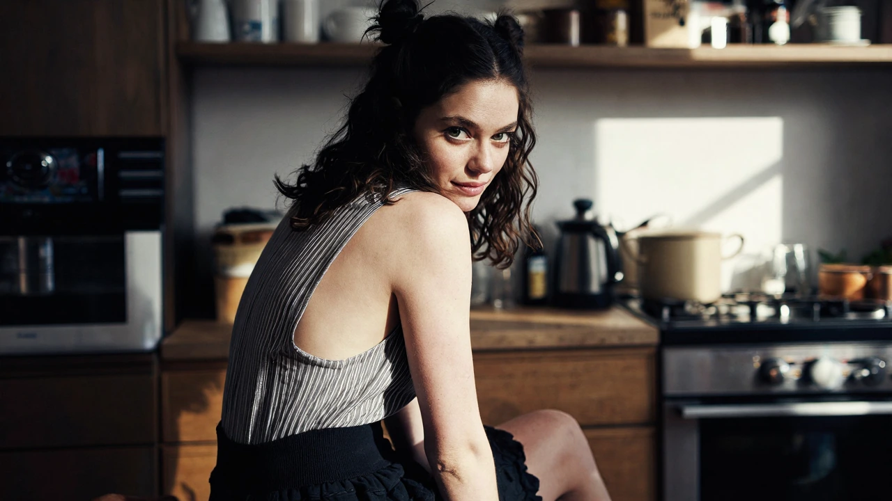 Young woman with dark wavy hair in twin buns, pale skin with freckles, green eyes, playful smile, seated in sunlit kitchen, pinstriped open-back top, 