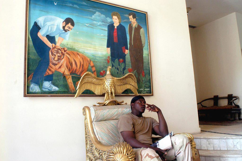 a US soldier rests in what used to be Saddam’s chair under a painting of Uday playing with a tiger,…